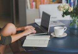 Female freelancer using laptop with coffee at home office desk, surrounded by roses and a planner.