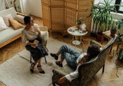 Two women engaged in a psychotherapy session in a warm, inviting interior with plants and natural lighting.